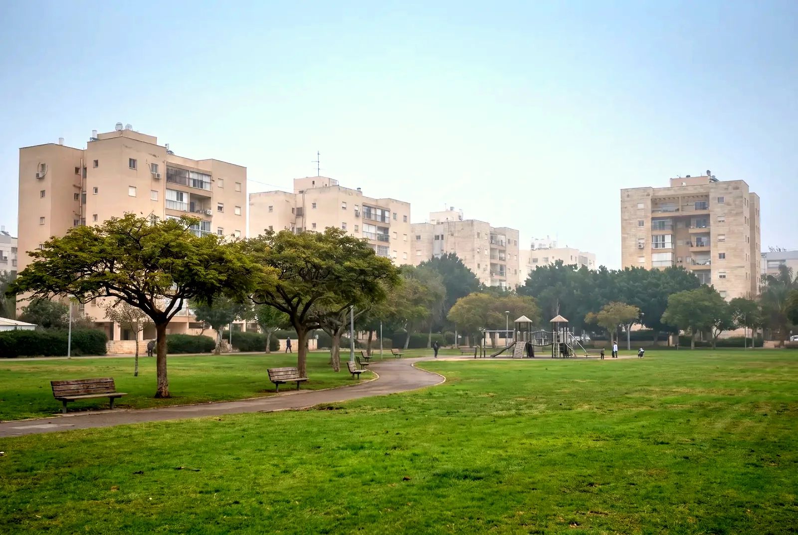 הדמיה: גן שכונתי במבט מרחוק, עם דשא, שבילים ובנייני מגורים מאחורי העצים