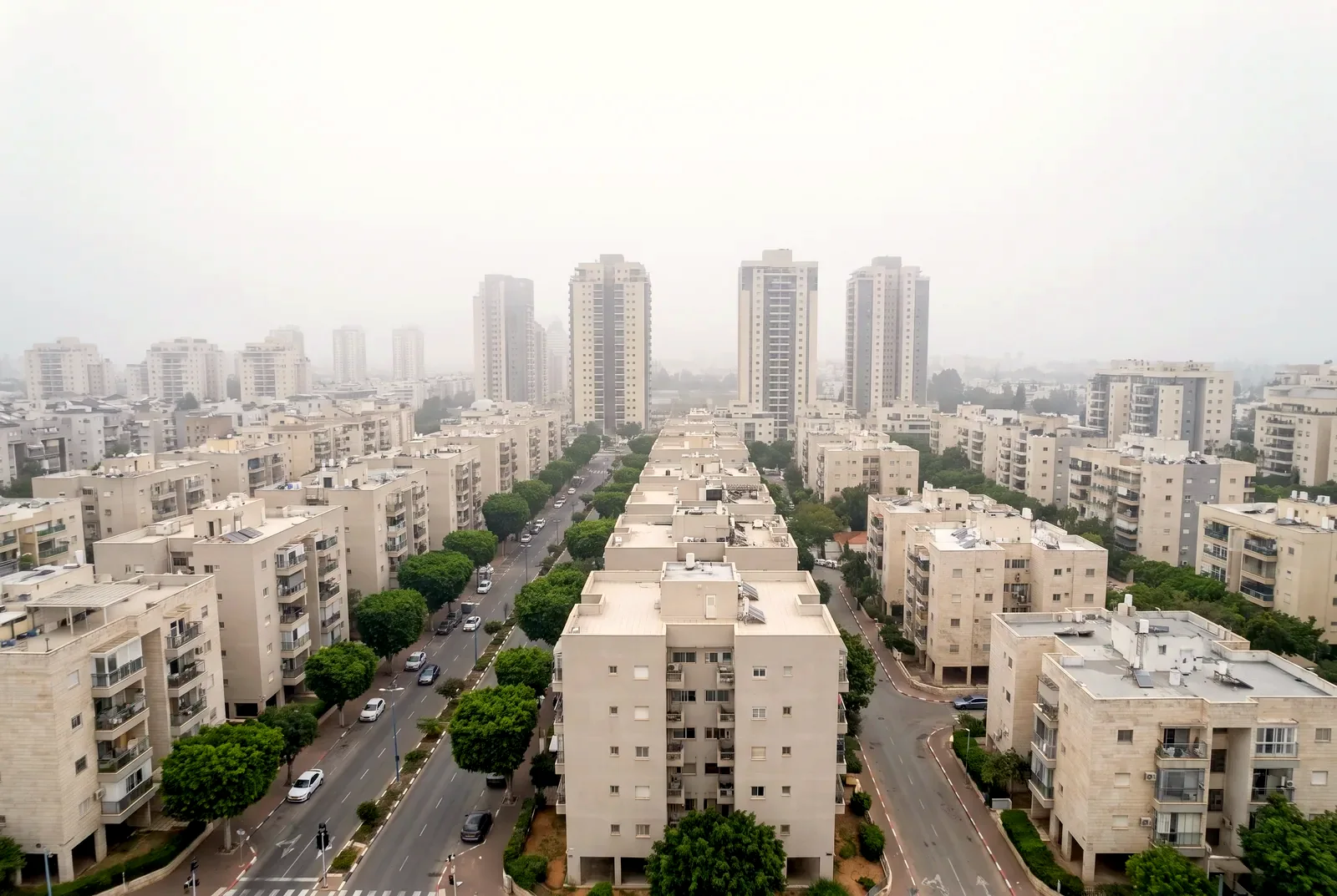 הדמיה: מבט מהאוויר באור מעונן רך על שכונות מגורים עם רחובות מוצלים ומגדלי מגורים באובך.
