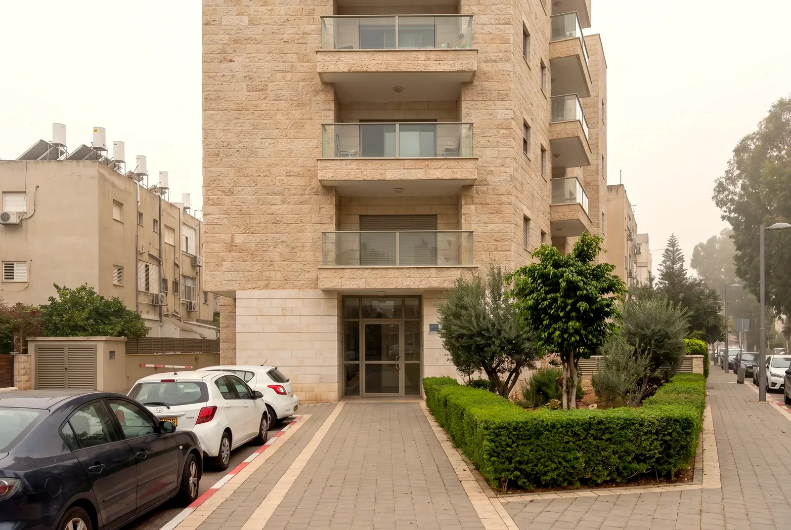 Illustration: the entrance to a new residential building clad in stone, with glass balconies and a landscaped garden, on a quiet city street