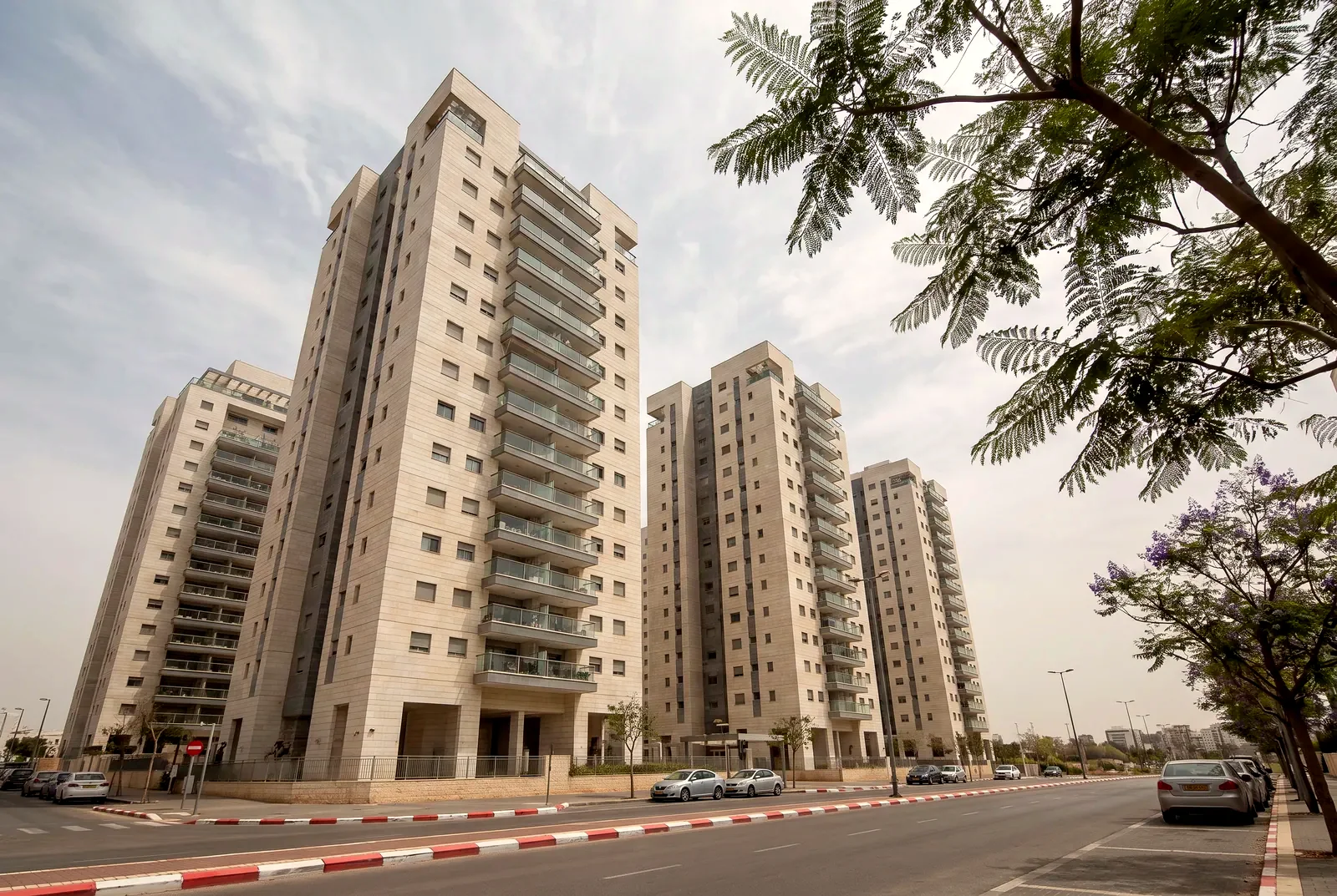 הדמיה: מבט מלמטה אל מגדלי מגורים חדשים מעל רחוב שקט עם עצי ג'קרנדה