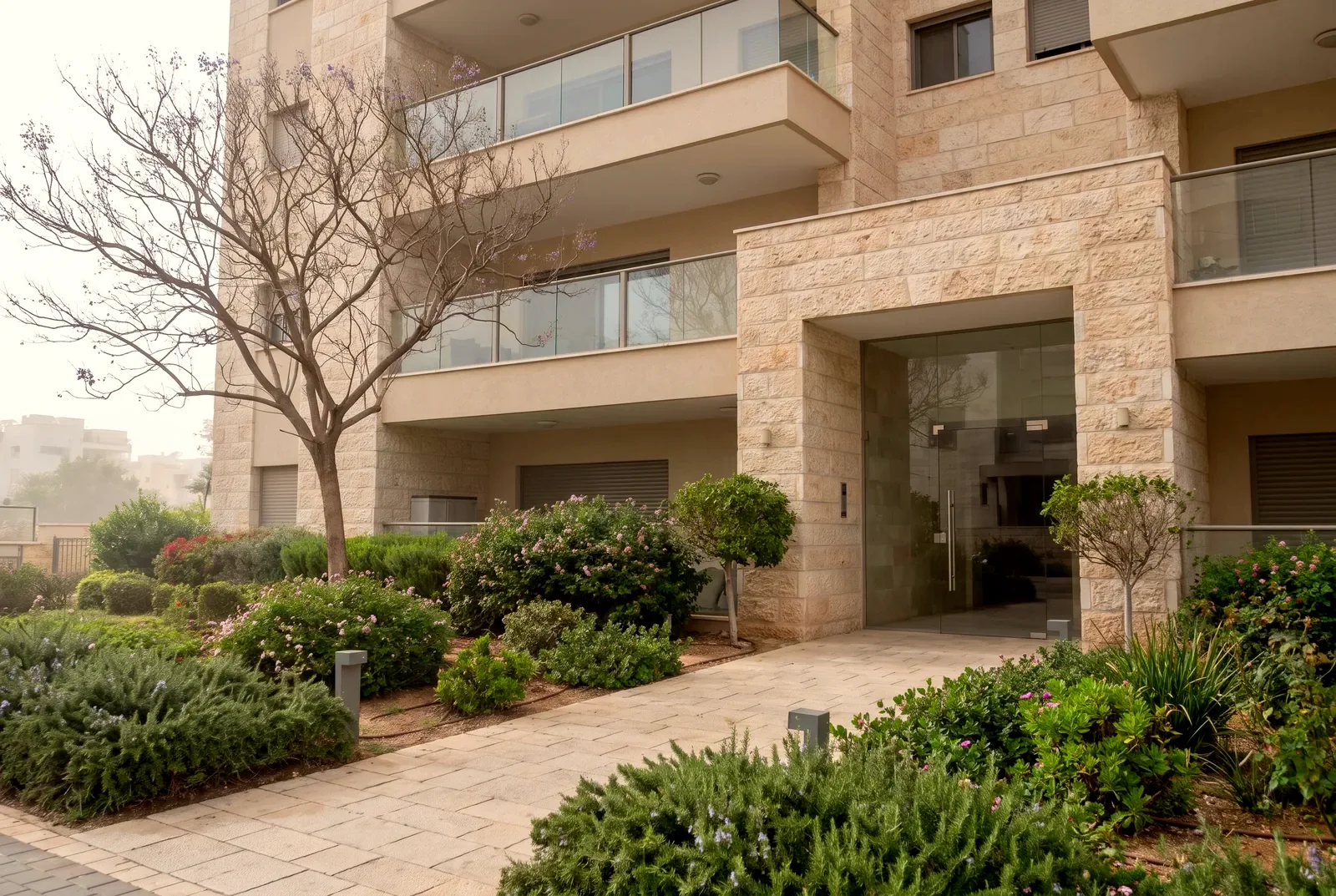 הדמיה: כניסה וגינה מטופחת של בניין מגורים חדש מחופה אבן עם מרפסות זכוכית.