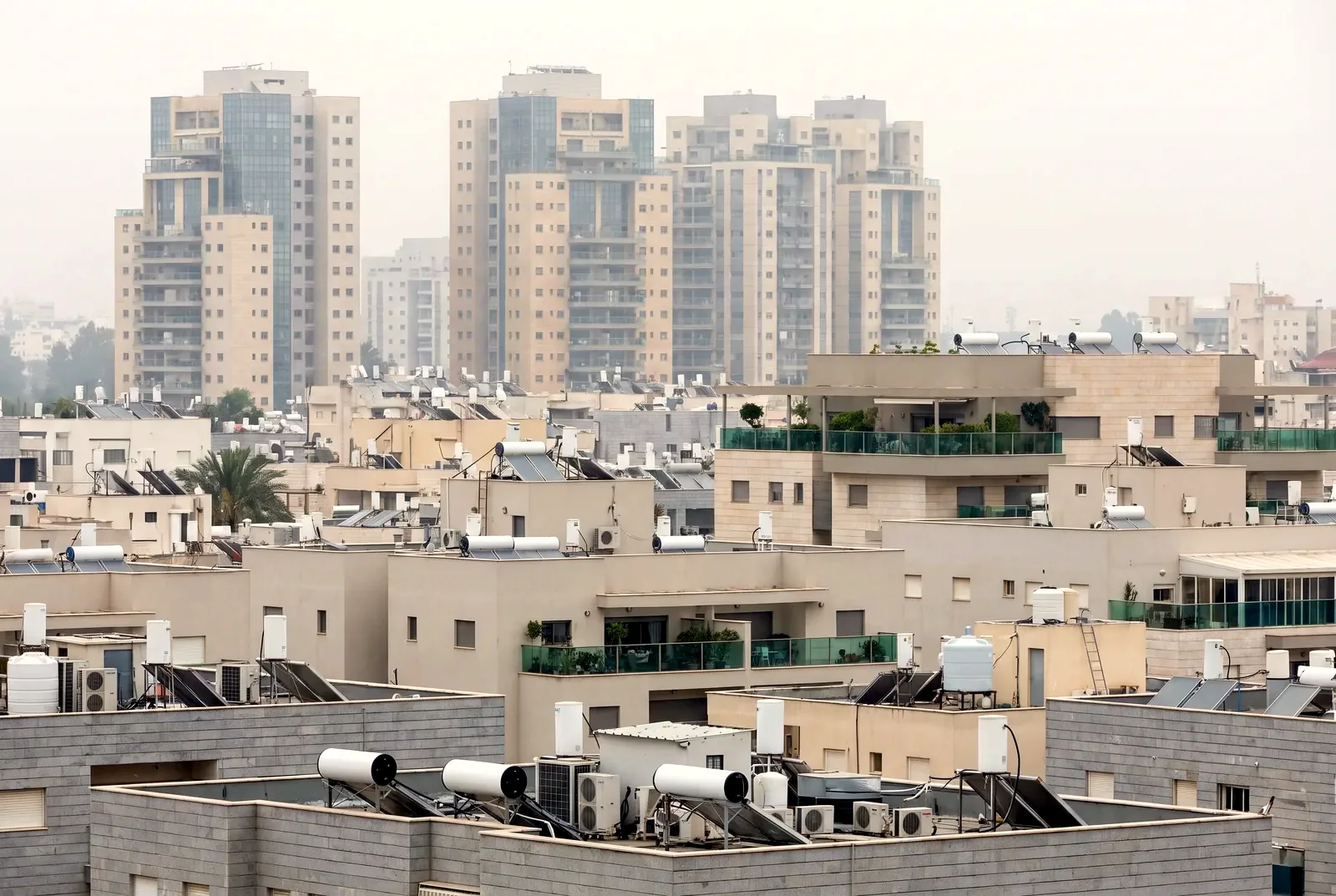 הדמיה: גגות שטוחים של בנייני דירות עם דודי שמש ומזגנים, ומגדלי מגורים באובך ברקע.