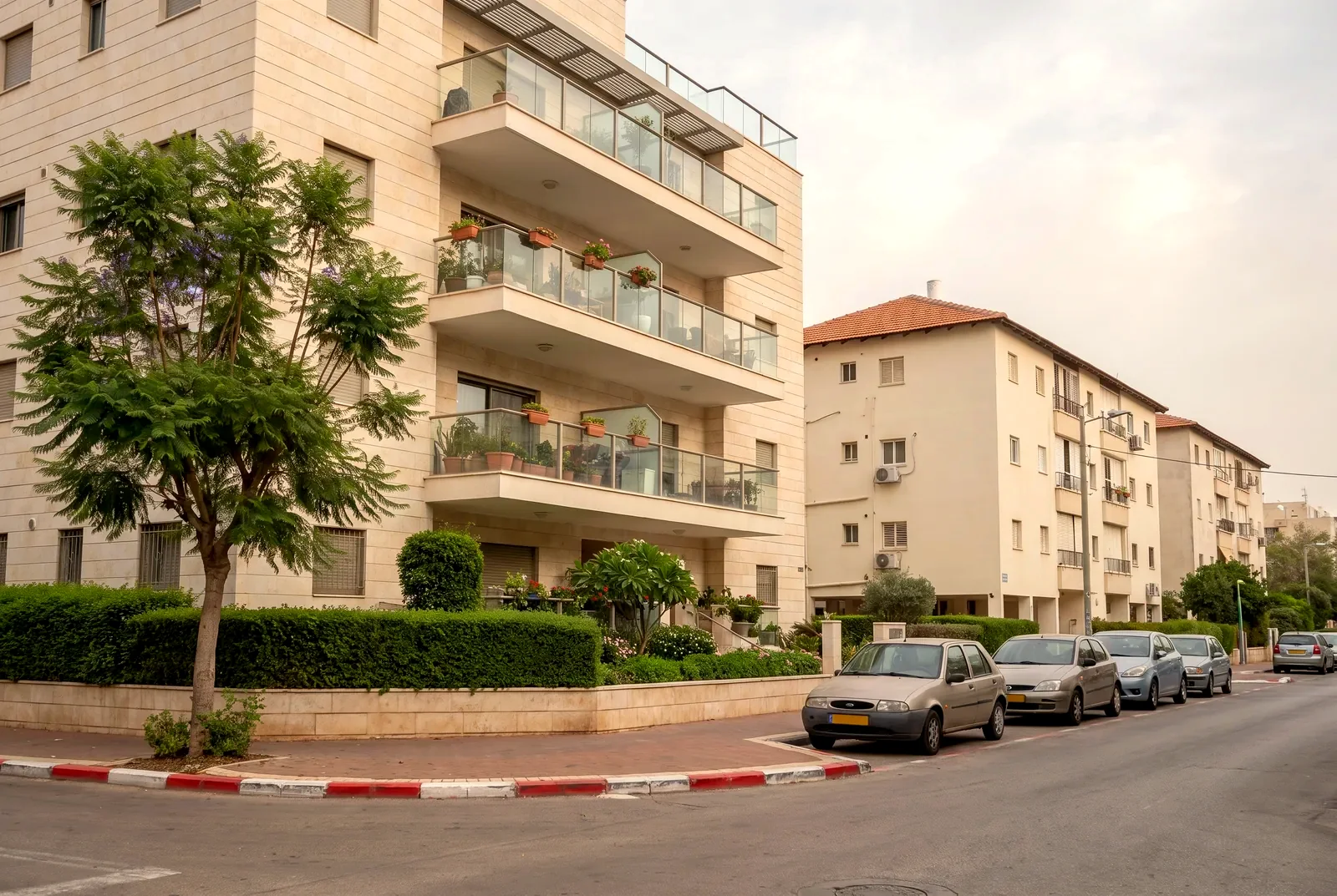 הדמיה: פינת רחוב שקט עם בניין מגורים חדש מחופה אבן, גינה קטנה ועץ ג'קרנדה.