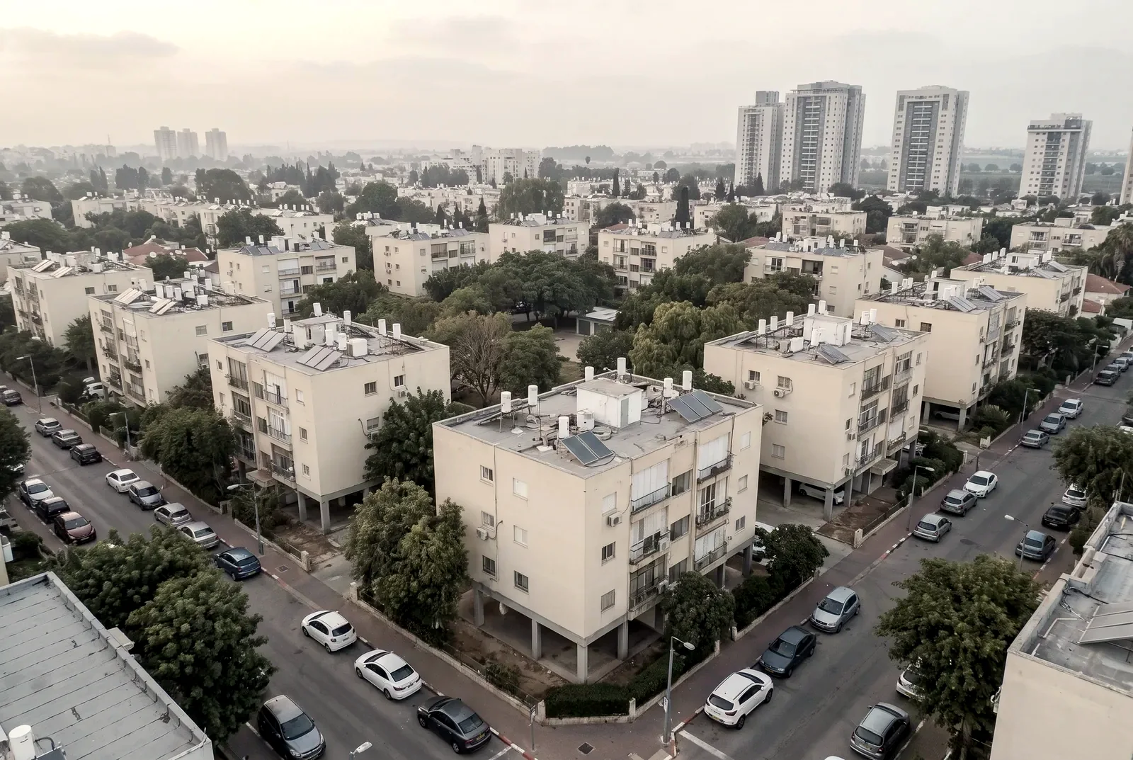 הדמיה: מבט אווירי על שכונה ותיקה ומסודרת עם בנייני עמודים ועצים, ומגדלים חדשים באופק
