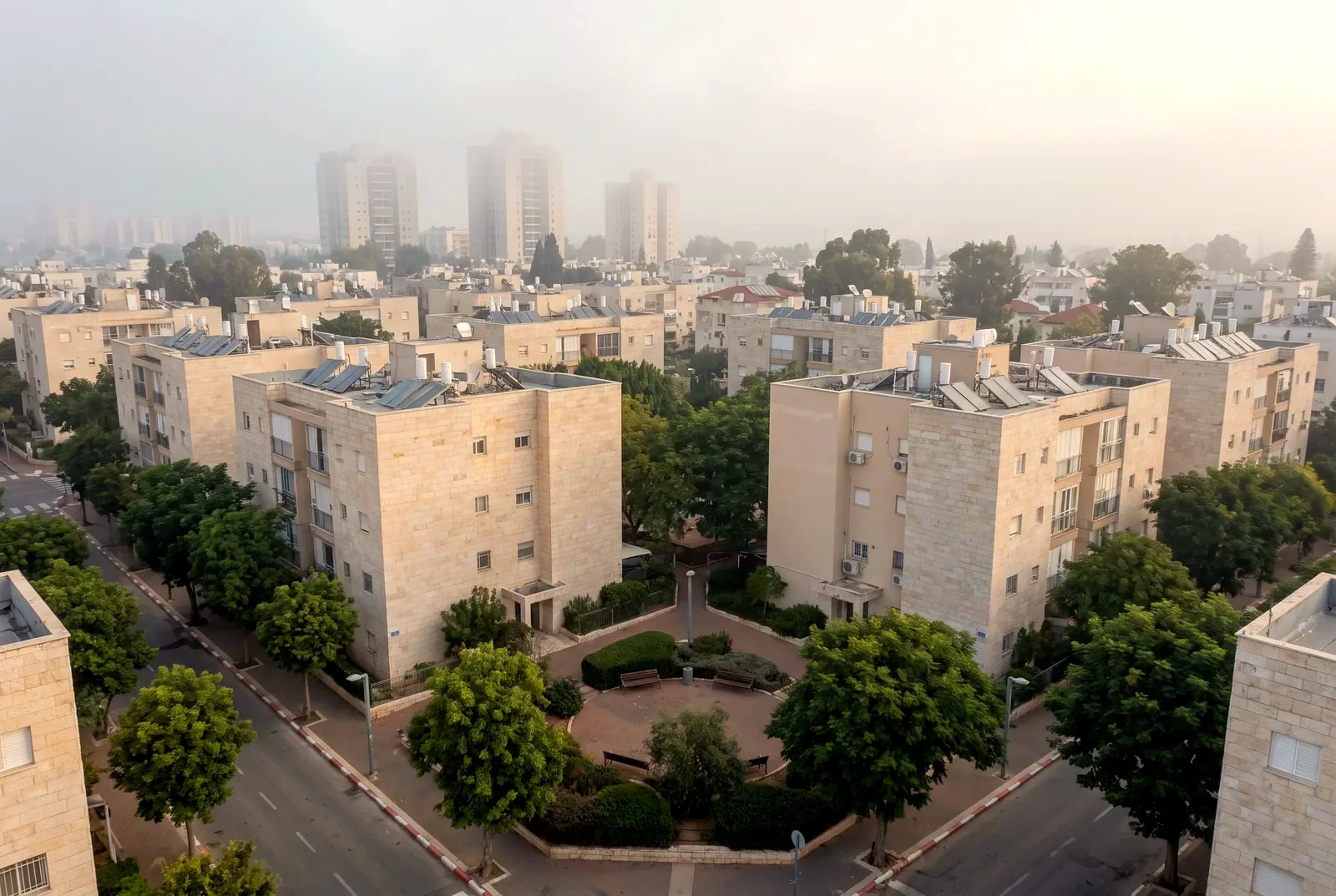 הדמיה: מבט רחפן נמוך על שכונה רגועה של בנייני מגורים עם חצרות ירוקות ומגדלים רחוקים