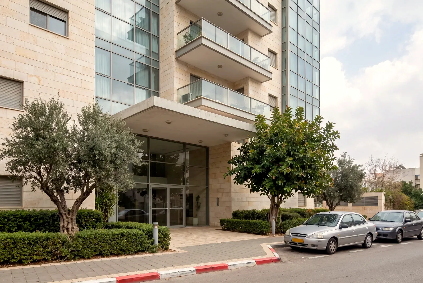 הדמיה: כניסה לבניין מגורים חדש ומטופח עם גינה מסודרת, עצי זית ופיקוס ומרפסות זכוכית