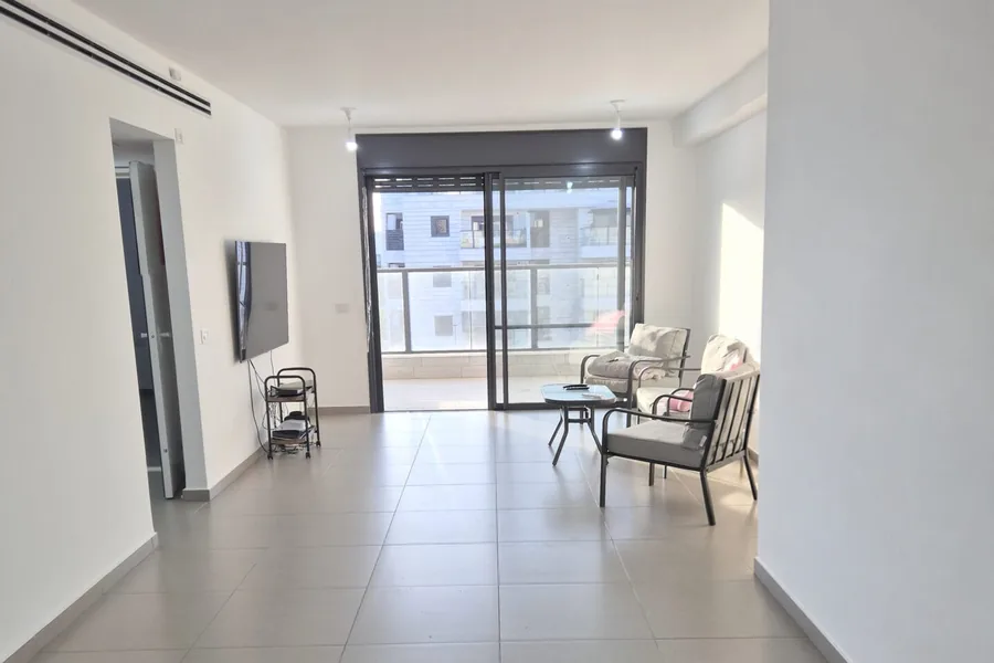 סלון בדירה ברחוב אלי ויזל 5 בראש העין: ריצוף בהיר, מסך על הקיר, כורסאות ויציאה למרפסת