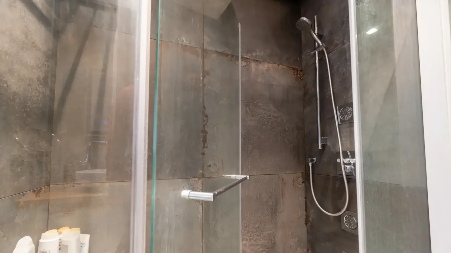 מקלחון זכוכית בציפוי אריחים אפור כהה, עם ראש מקלחת ומוט.