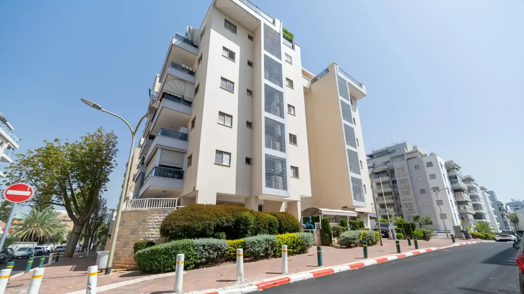חזית הבניין מהרחוב: בניין מגורים בהיר מרובה קומות עם מרפסות, שיחים ומדרכה, שמיים כחולים.