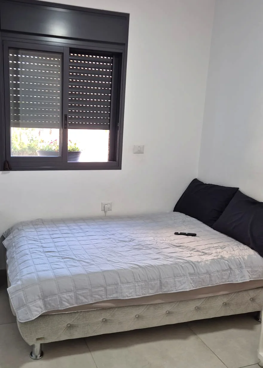 חדר שינה נוסף: מיטה עם כריות כהות מתחת לחלון עם תריס גלילה, קירות לבנים ורצפת אריחים בהירה.